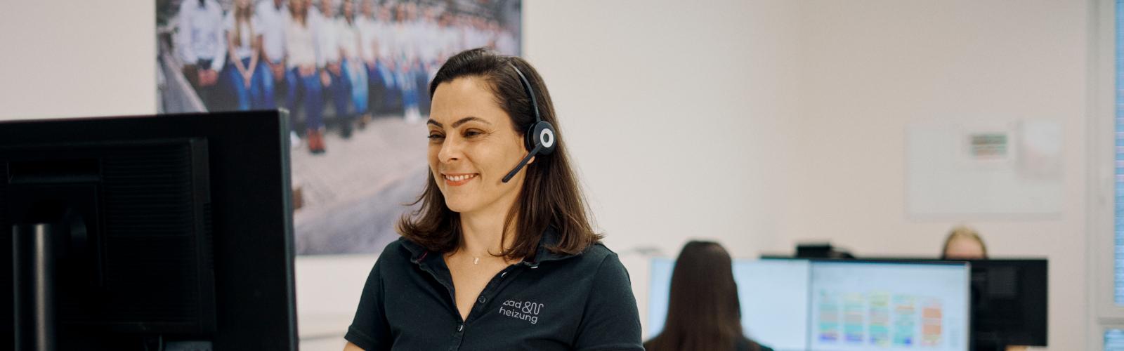 Eine Frau mit einem Headset sitzt lächelnd an einem Schreibtisch in einem modernen Büro.