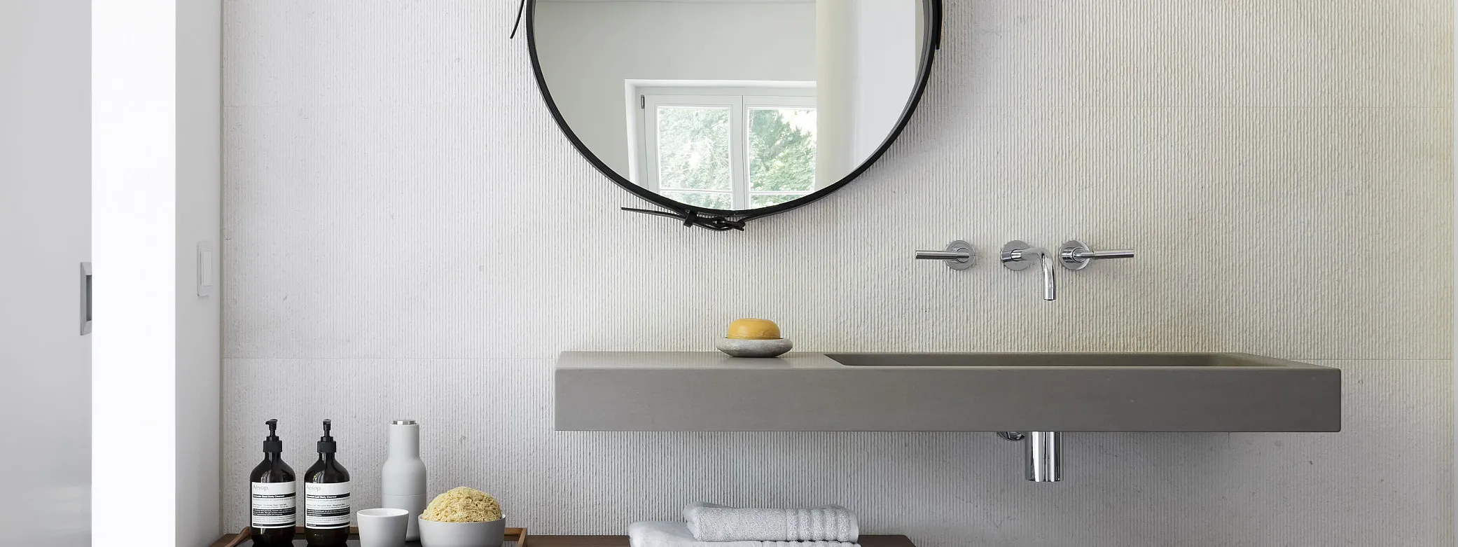Minimalistisch gestaltetes Badezimmer mit runden Wandspiegel, grauem Waschbecken und einer Ablagefläche für Pflegeprodukte und Handtücher.