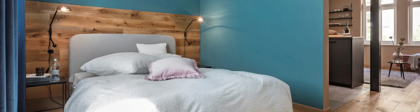 Polsterbett mit heller Bettwäsche und Holzrückwand, flankiert von zwei Leselampen, blauer Wandanstrich und Blick in angrenzendes Zimmer mit Sitzbereich.