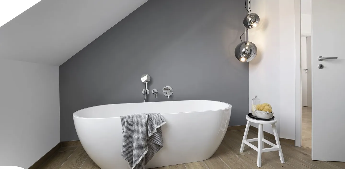 Badezimmer unter einer Dachschräge, dominiert von einer weißen, ovalen Badewanne vor einer dunkelgrauen Wand.