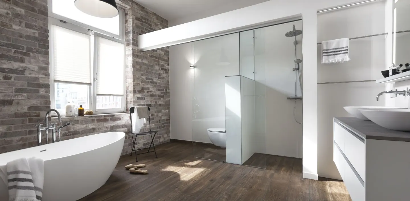 Geräumiges Badezimmer mit freistehender Badewanne vor einer Backsteinwand und Duschbereich mit Glasabtrennung. Ein Holzfußboden ergänzt das Design.