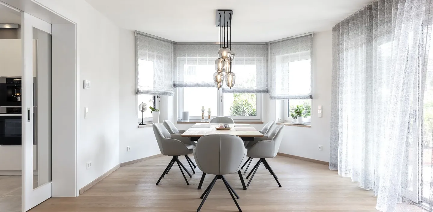 Esszimmer mit Holzboden, einem rechteckigen Holztisch, umgeben von sechs grauen Polsterstühlen mit schwarzen Beinen. 