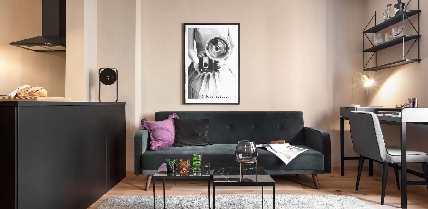 Wohnzimmer mit dunkelgrünem Sofa, zwei Couchtischen auf grauem Teppich, gerahmtem Bild an der Wand und Schreibtisch mit Regal im Hintergrund.