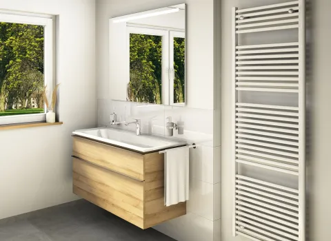 Geräumiges Badezimmer mit einem Holz-Waschtisch, Spiegel und einem Fenster mit Blick ins Grüne. Rechts an der Wand befindet sich ein vertikaler Handtuchheizkörper.