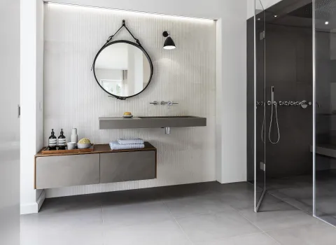 Badezimmer mit einem runden Wandspiegel, grauem Waschbecken und separater Duschkabine aus Glas.