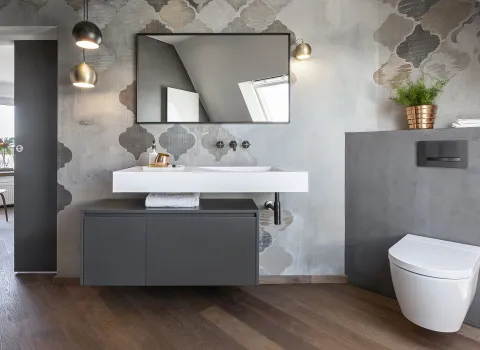 Badezimmer mit einem weißen Waschtisch auf einem schwarzen Unterschrank, einem rechteckigen Spiegel und einer gemusterten Akzentwand. Zwei Hängelampen beleuchten den Waschbereich, rechts steht eine weiße Toilette.