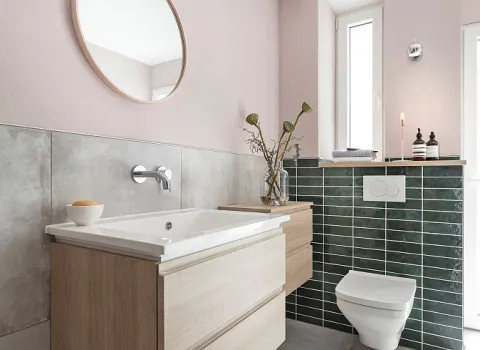 Helles Badezimmer mit Holz-Waschtisch, rundem Spiegel, wandhängender Toilette und grüner Fliesenverkleidung, daneben Fenster und Deko.