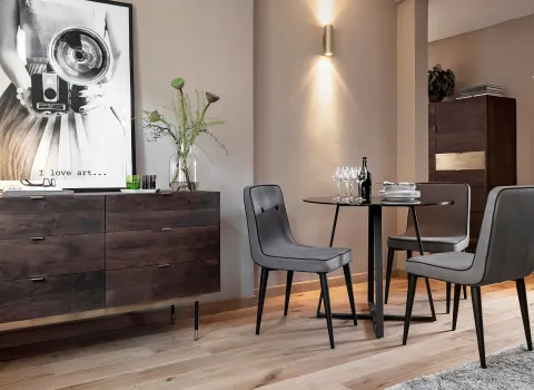 Essbereich mit rundem Tisch und zwei grauen Stühlen, daneben eine Kommode mit Dekor und großem gerahmten Foto, beleuchtet durch Wandleuchte.