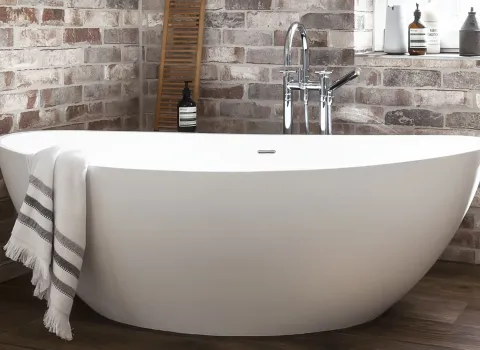 Weiße, freistehende Badewanne vor einer Backsteinwand, mit einem Handtuch über den Rand gelegt.