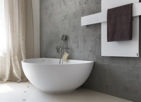Badewanne mit asymmetrischem Design vor grauer Fliesenwand. Links fällt Tageslicht durch einen Vorhang, rechts ist ein weißer Handtuchwärmer mit Handtuch sichtbar.