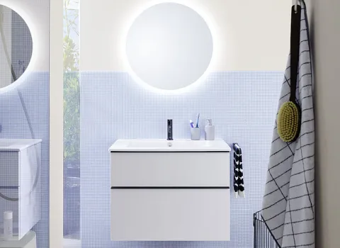 Das Badezimmer der Eqio-Kollektion präsentiert sich mit einem schlichten weißen Waschtisch und einem runden LED-Spiegel, der sanftes Licht abgibt.