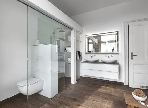 Helles Badezimmer mit Dusch- und WC-Bereich hinter einer Glaswand sowie einem Doppelwaschplatz mit großem Spiegel. Der Boden ist aus dunklem Holz.