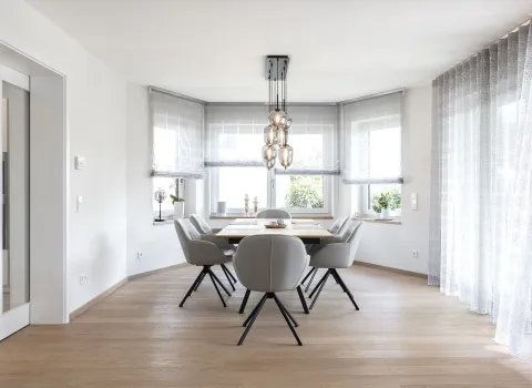 Esszimmer mit Holzboden, einem rechteckigen Holztisch, umgeben von sechs grauen Polsterstühlen mit schwarzen Beinen. 