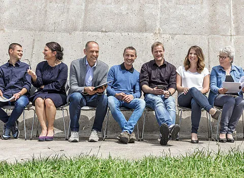 SiebenMitarbeiter der Kirchgäßner GmbH sitzen in Business-Casual-Kleidung vor einer Mauer und lächeln.