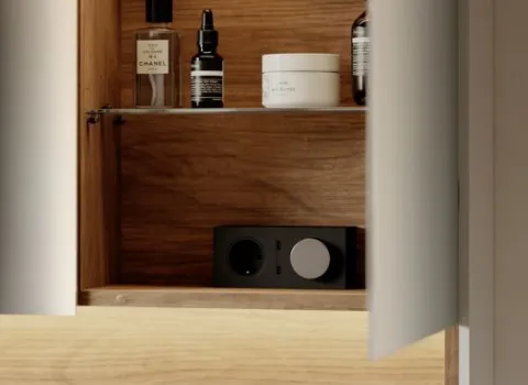 Ein geöffneter Spiegelschrank mit edlem Holzdekor zeigt sorgfältig arrangierte Pflegeprodukte und ein integriertes Steckdosenmodul im unteren Fach.