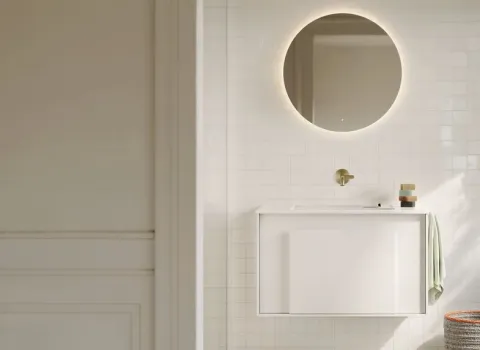 Ein heller Gästebad Waschtisch mit weißer Front und goldfarbener Armatur ist durch eine offene Tür sichtbar.