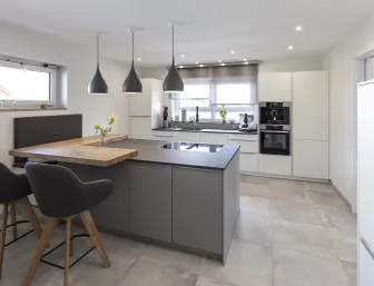 Helle Küche mit einer grauen Kücheninsel, die einen Holzaufsatz als Frühstückstheke und zwei Barhocker in dunklem Grau bietet.