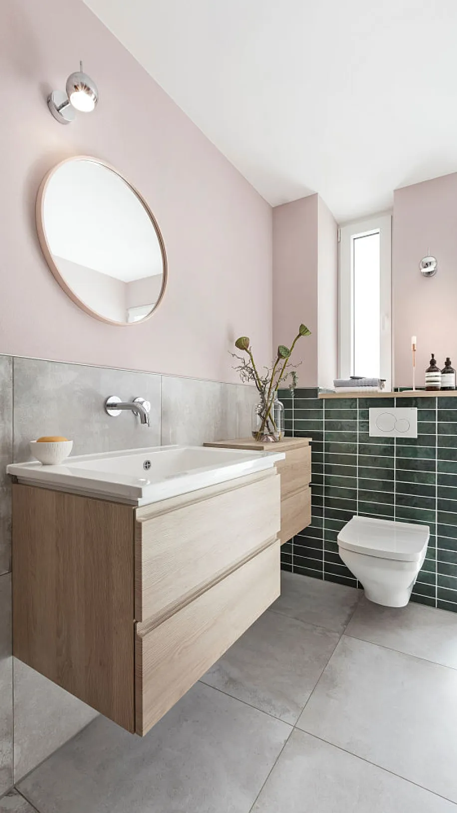 Helles Badezimmer mit Holz-Waschtisch, rundem Spiegel, wandhängender Toilette und grüner Fliesenverkleidung, daneben Fenster und Deko.