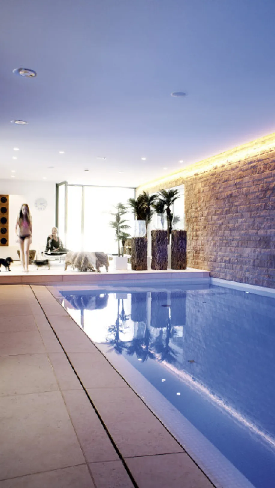 Großer Innenpool mit blauen Wasserreflexionen, umgeben von heller Beleuchtung und einer rustikalen Steinwand.