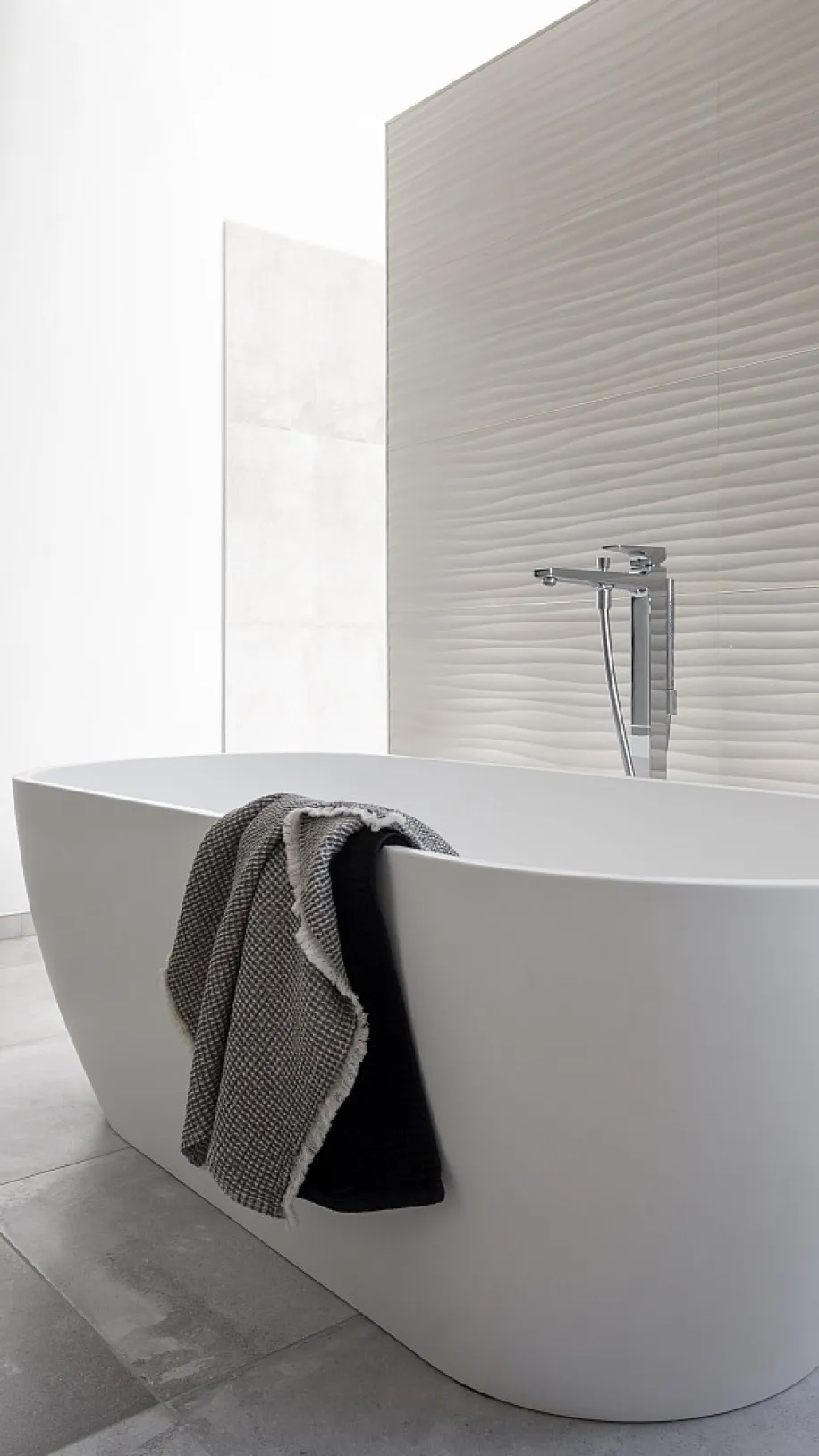 Helle Badewannenecke mit minimalistischer Wanne und einem Handtuch über dem Rand. Die strukturierte Wand und der kleine Beistelltisch betonen das moderne Design.