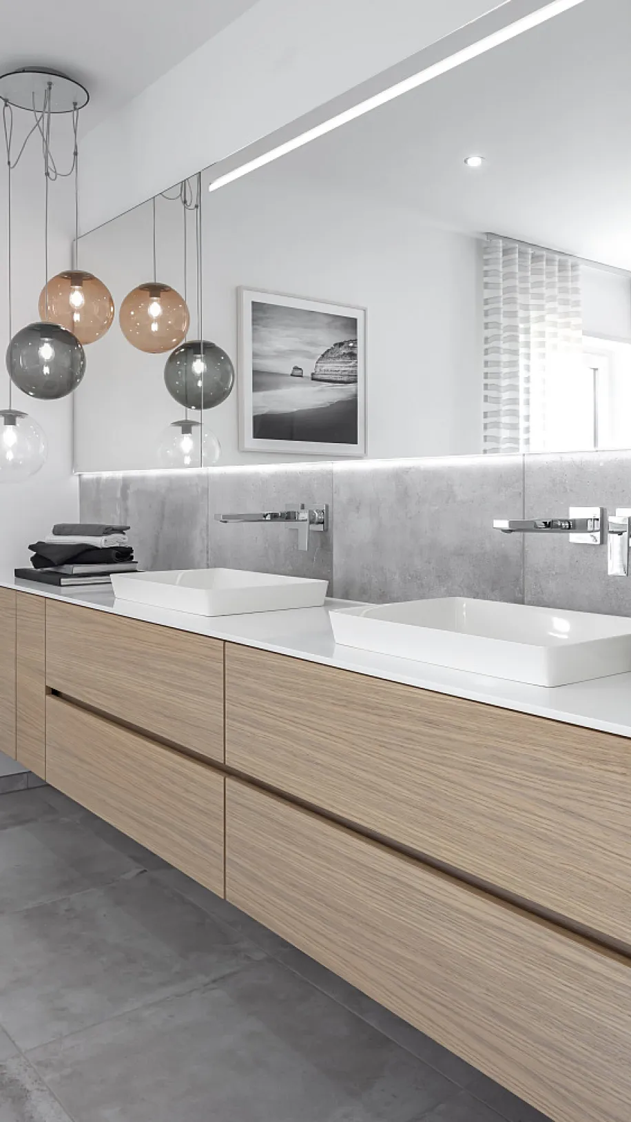 Doppelwaschtisch aus Holz mit zwei Waschbecken und großem Wandspiegel. Links hängen runde Lampen von der Decke, an der Wand sind zwei Schwarz-Weiß-Bilder.