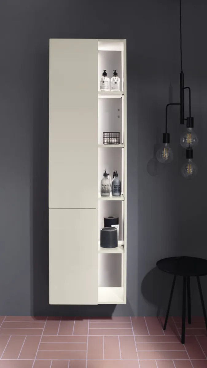 Der schlanke Burgbad Badu Hochschrank im Badezimmer ist halb geöffnet und zeigt beleuchtete Fächer, die Pflegeprodukte wie Seifenspender und Kerzen beinhalten.