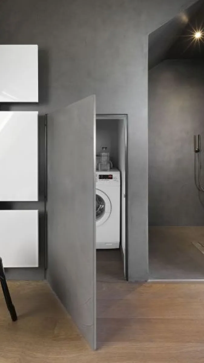 In grau gehaltenes Badezimmer mit Holzboden und halboffener Wandtür, hinter der eine Waschmaschine untergebracht ist.