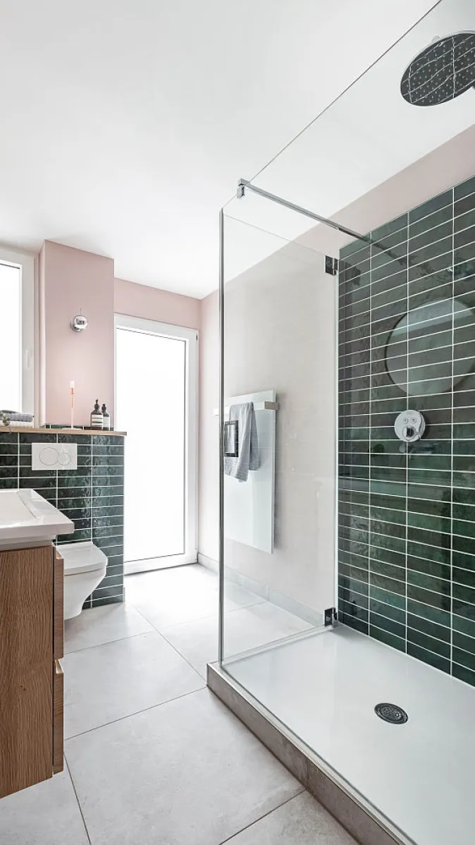 Badezimmer mit bodengleicher Glasdusche, grün gefliester Duschwand, hellem Boden und einer Toilette neben einem kleinen Fenster.