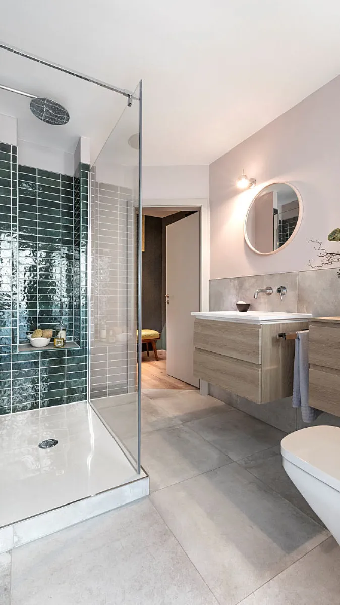 Helles Badezimmer mit bodengleicher Dusche, grün gefliester Duschwand, Holzwaschtisch und runden Spiegelakzenten.