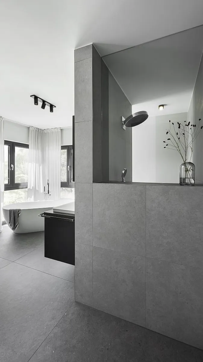 Badezimmer mit grauer Fliesenwand und abgetrennter Duschkabine. Im Hintergrund sind eine Badewanne und große Fenster mit weißen Vorhängen zu sehen.
