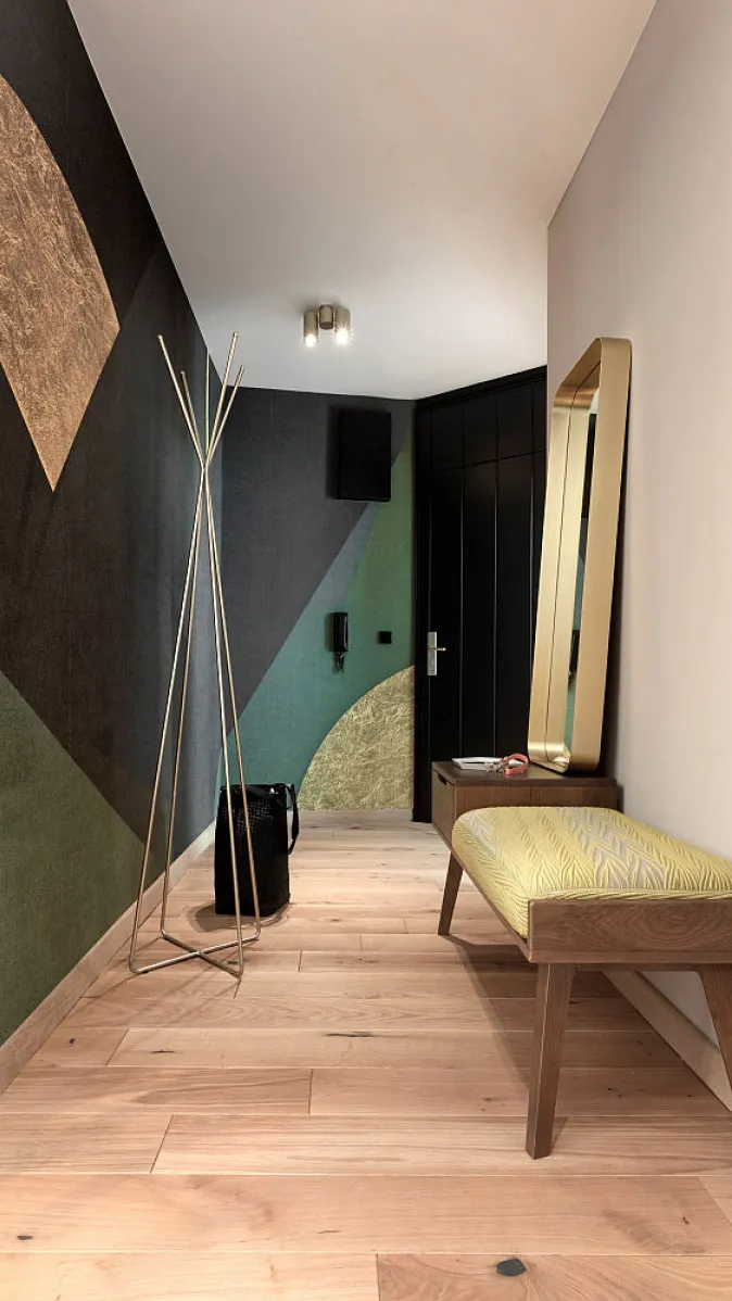 Eingangsbereich mit gemusterter Wand, heller Holzbank mit Polster, schmalem Spiegel in Gold und filigranem Garderobenständer.