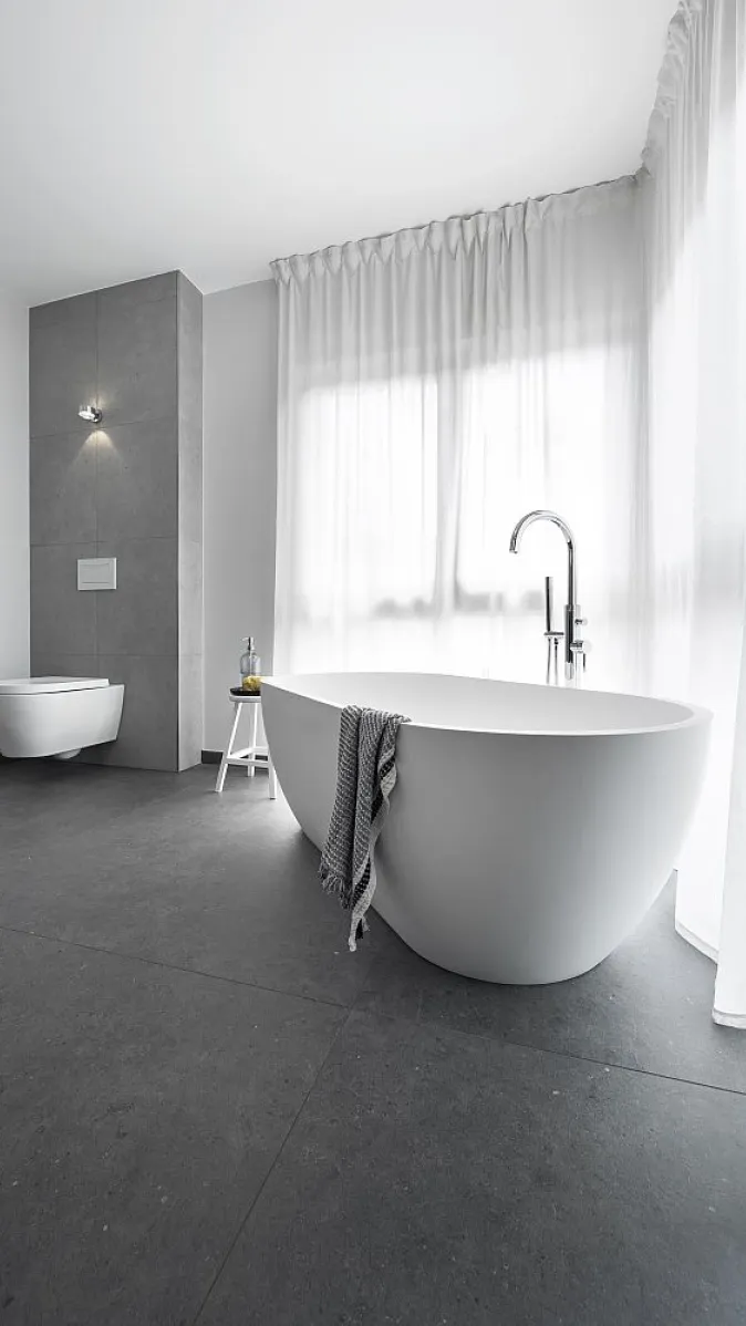 Freistehende Badewanne in einem modernen, hellen Badezimmer mit grauem Fliesenboden und einem großen Fenster mit weißen Vorhängen. Ein Handtuch hängt über dem Badewannenrand.