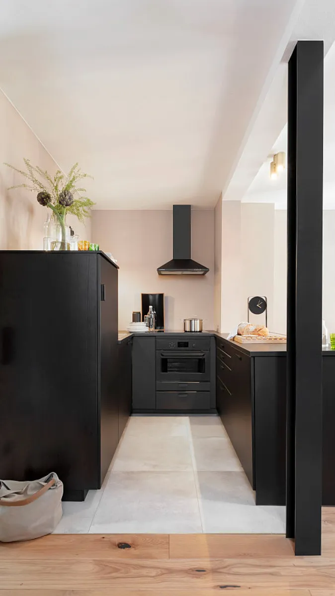 Schmale Küche mit schwarzen Schränken, schwarzem Backofen und Dunstabzugshaube, dazu beige Wände und hellgrauer Fliesenboden.