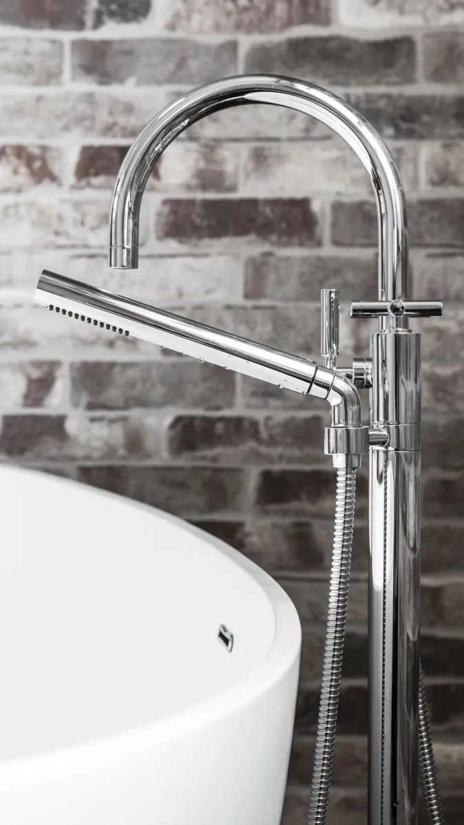 Freistehende Armatur mit glänzendem Wasserhahn und Handbrause neben einer Badewanne.