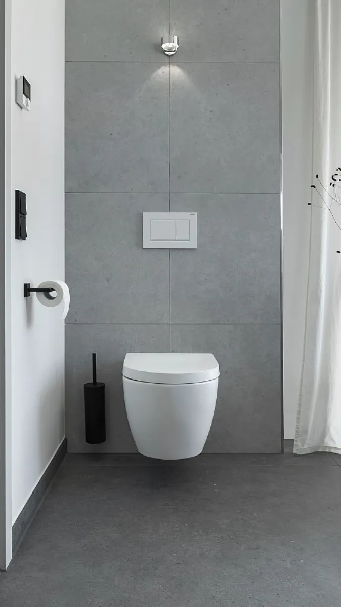 Modernes Badezimmer mit wandhängender Toilette vor einer grauen Fliesenwand. Daneben steht ein kleiner Tisch mit Vase und Trockenblumen.