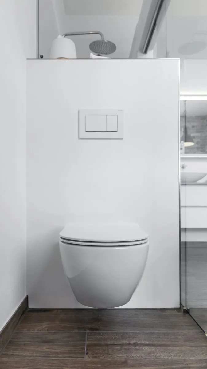 Weiße Toilette mit Unterputz-Wandspülung vor einer weißen Trennwand, die den Duschbereich abgrenzt.