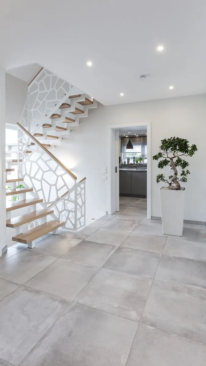 Ein tageslichtheller Eingangsbereich mit einer offenen Treppe aus hellen Holzstufen und einem auffälligen weißen Geländer in netzartiger Form.