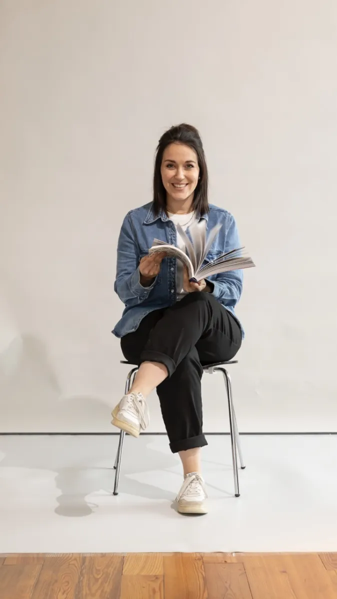 Mitarbeiterin Jasmin Englert sitzt lächelnd mit überschlagenen Beinen auf einem Stuhl und hält einen geöffneten Katalog in den Händen.