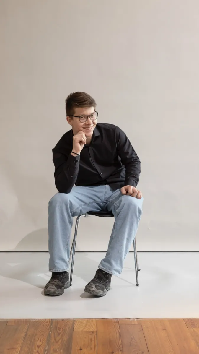 Lächelnder Mitarbeiter mit Brille sitzt locker auf einem Stuhl und stützt das Kinn auf eine Hand.