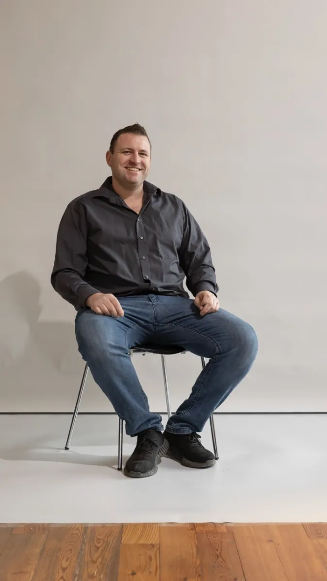 Lächelnder Mitarbeiter in schwarzem Hemd und Jeans sitzt entspannt auf einem Stuhl vor hellem Hintergrund.