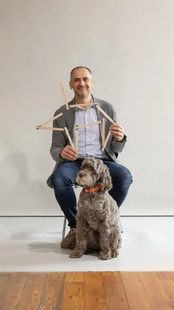 Hans Kirchgäßner sitzt lächelnd auf einem Stuhl, hält mehrere Zollstöcke in den Händen und wird von einem Hund mit orangefarbenem Halsband begleitet.