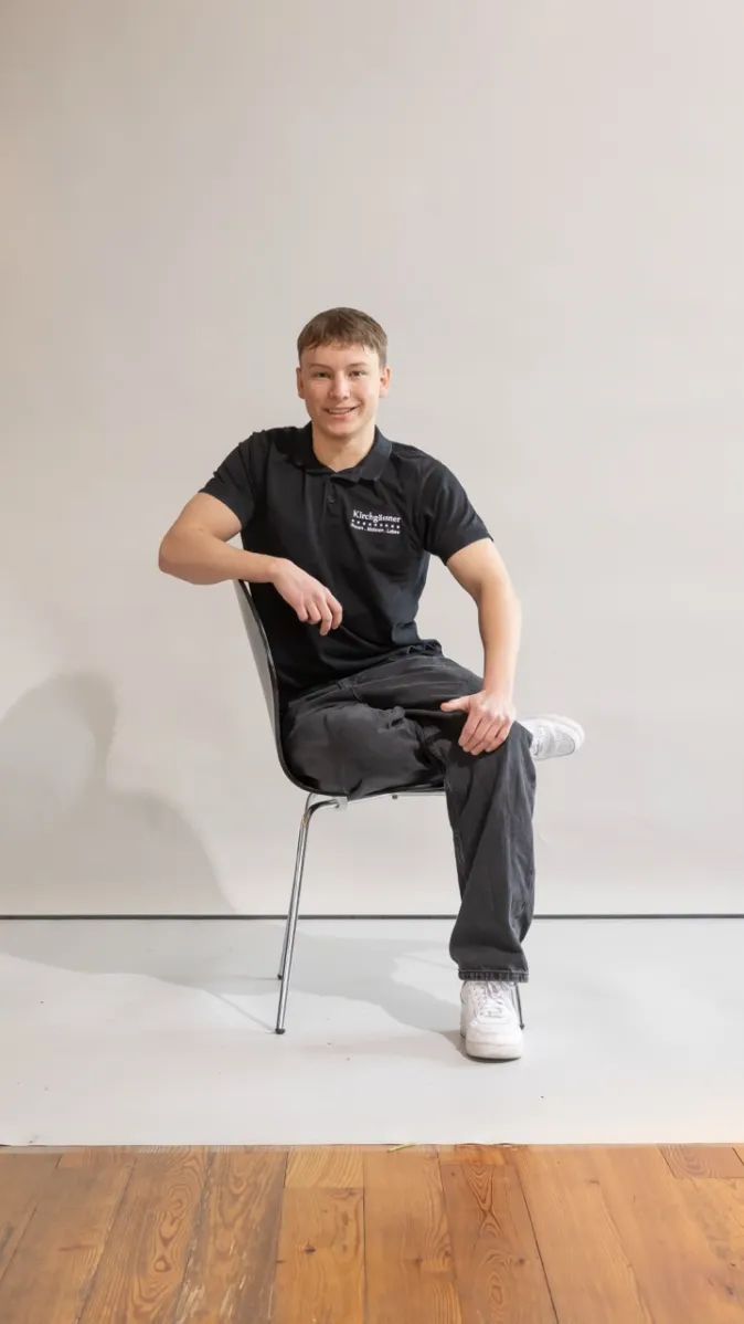 Mitarbeiter Theo sitzt locker mit überschlagenen Beinen auf einem Stuhl und lächelt in die Kamera.