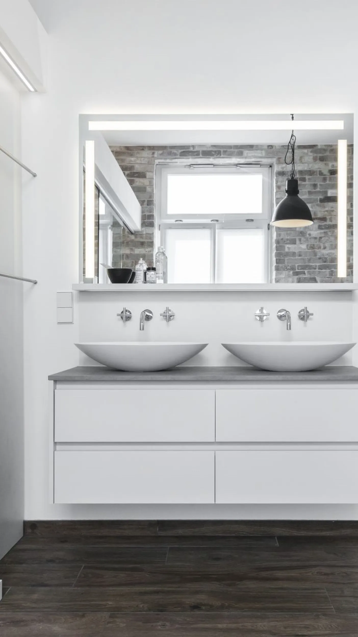 Doppelwaschbecken mit rundem Aufsatz auf einem schwebenden weißen Waschtisch. Der große Spiegel dahinter reflektiert ein Fenster und eine Backsteinwand.