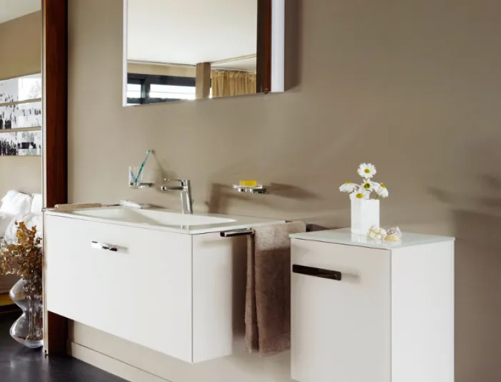 Ein modernes Badezimmer mit einem weißen Waschbecken, das an einer glatten, beige gestrichenen Wand montiert ist. Ein großer Spiegel hängt über dem Waschbecken, das von minimalistischen Schränken flankiert wird, auf denen eine Vase mit weißen Gänseblümchen steht. Die Oberflächen sind makellos und spiegeln die professionelle Arbeit von Malern und Trockenbauern wider, die eine elegante und ruhige Atmosphäre geschaffen haben.