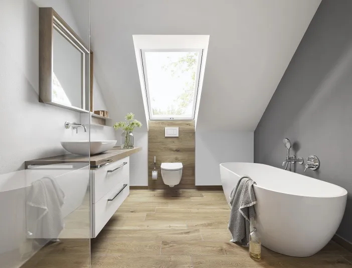 Helles Dachbadezimmer mit einer freistehenden weißen Badewanne und einer wandmontierten Toilette unter einem großen Dachfenster, das viel natürliches Licht hereinlässt.