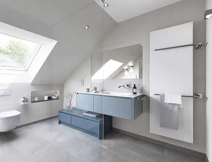 Helles Dachbad mit schwebendem Waschtisch, dunkelblauem Stauraum und großem Fenster. Handtuchheizkörper und graue Bodenfliesen schaffen minimalistischen Look.