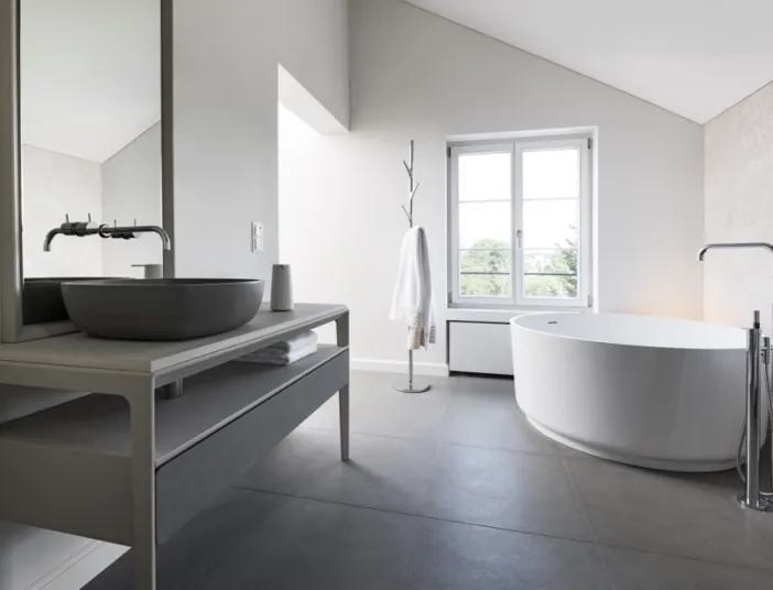 Badezimmer mit ovaler freistehender Badewanne neben einem großen Fenster. Links steht ein Waschtisch mit rundem Waschbecken und Wandarmaturen.