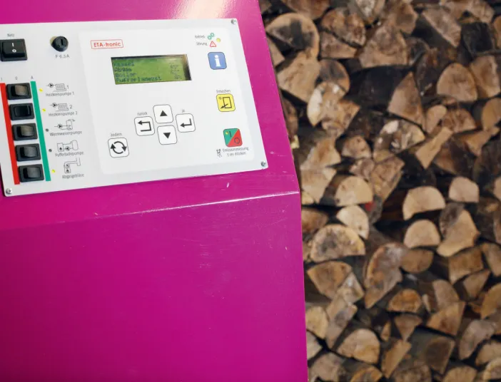 Rosa Heizungssteuerung mit Display und Tastenfeld, im Hintergrund dicht gestapeltes Feuerholz.