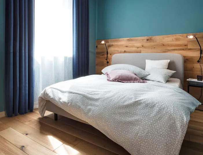 Doppelbett mit heller Bettwäsche vor einer Rückwand aus Holz, blauer Wandanstrich, links ein Fenster mit blauem Vorhang, rechts ein kleiner Nachttisch.